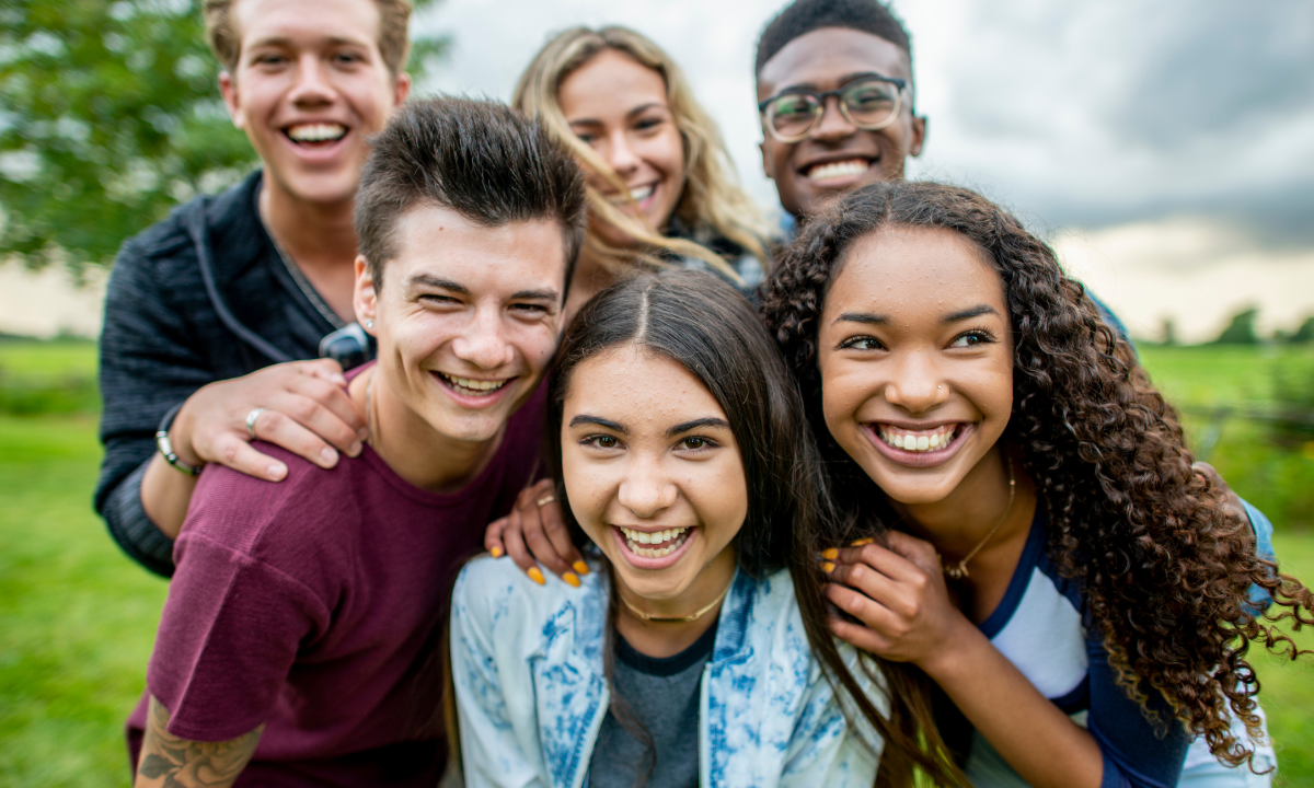 A multi-ethnic group of high school students are outdoors on a summer day. They are gathered together to take a selfie, and they are all smiling.